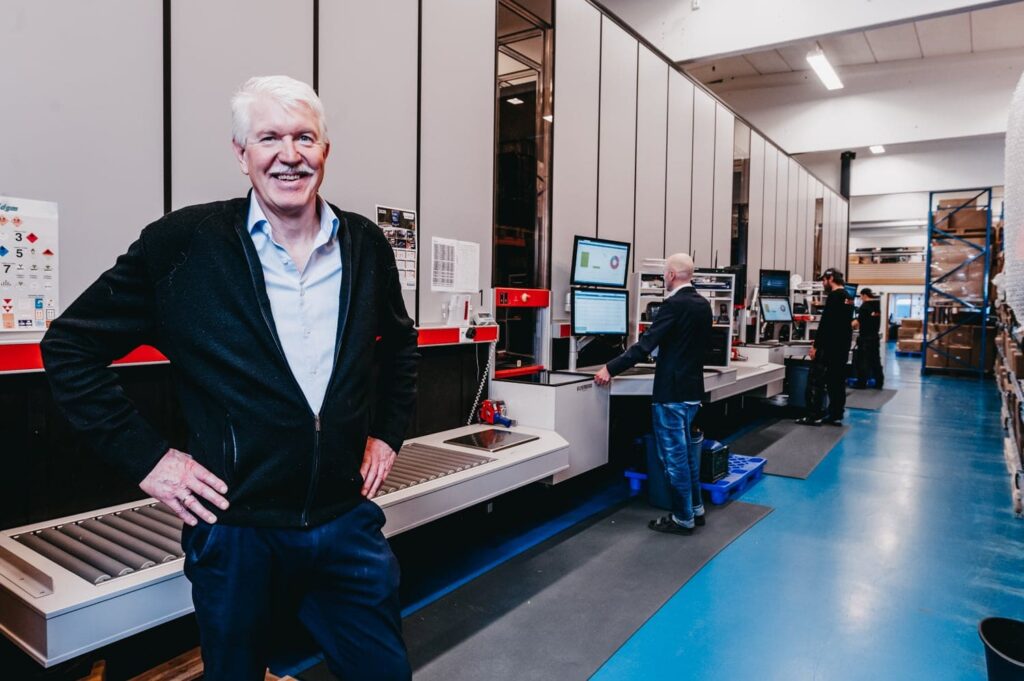 Managing director, Jan Kleven, smiles proudly to the camera in front of Elotecs AutoStore-solution with workers operating the ports in the background.