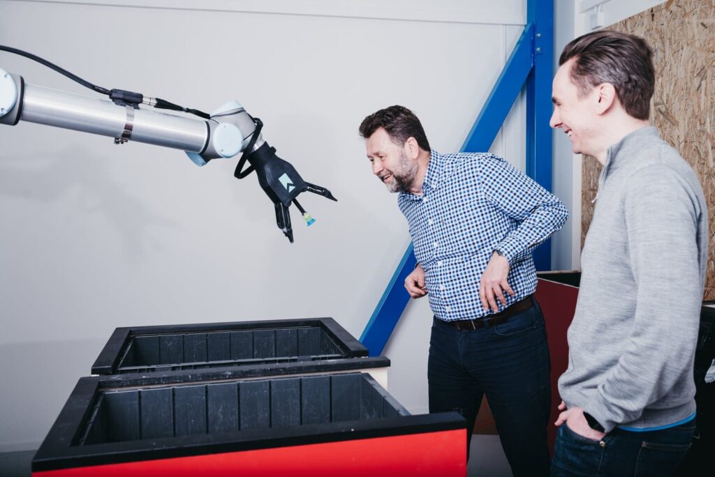 Two men standing by and observing a robotic arm while it grips for items.