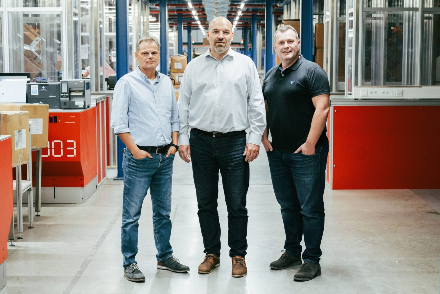 Three men standing next to each other at a warehouse, workstations in the background