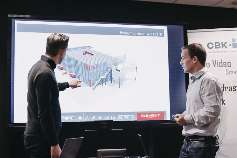 Two men talking and looking at a big screen portraying a 3d model of a warehouse