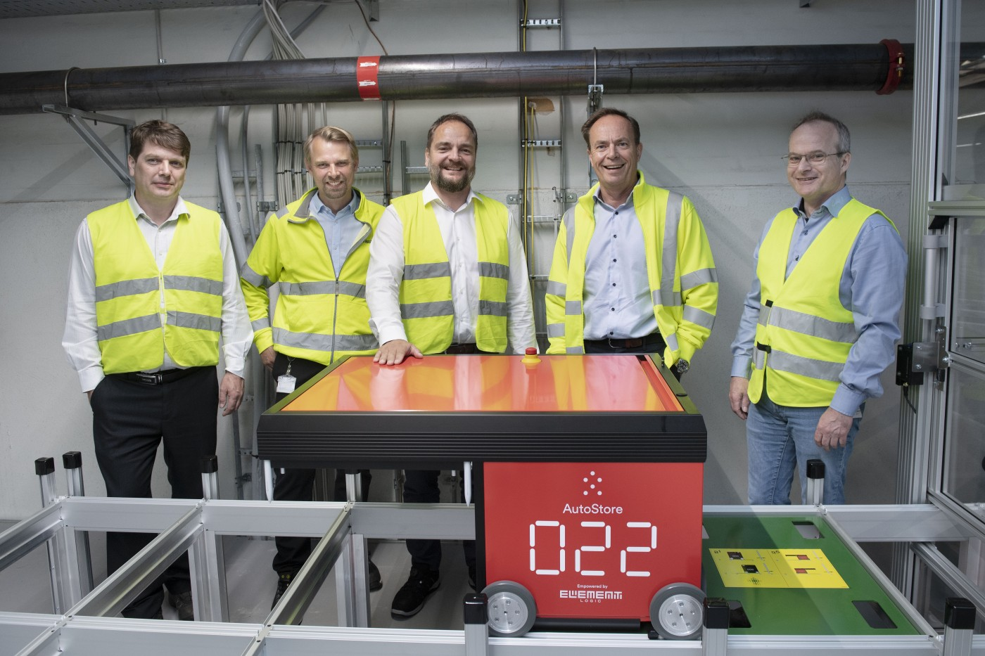 Five men in yellow vests standing behind an autostore robot