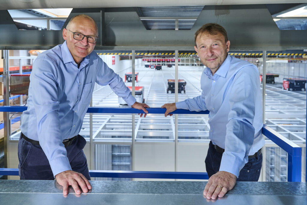 Dos hombres sonrientes, vestidos con camisas azules, se apoyan en una barandilla con un sistema AutoStore en funcionamiento al fondo. Robots de color negro y rojo se desplazan sobre una estructura de almacenamiento de rejillas.