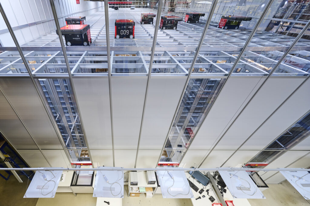 Vista aérea de una instalación de AutoStore en un centro de microfulfillment, donde se observan robots negros y rojos desplazándose sobre una estructura de almacenamiento de rejillas. Debajo se aprecian estaciones de trabajo con equipos electrónicos.