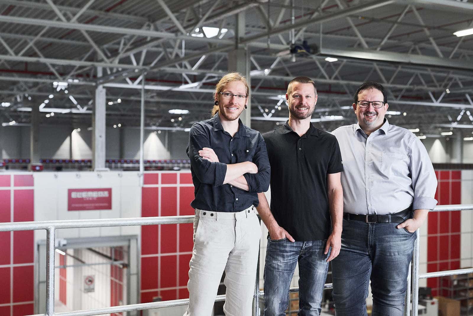 Tres directivos de Bergfreunde y Element Logic posan en un almacén automatizado con AutoStore en el fondo.