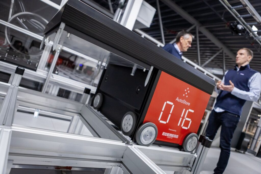 Imagen de un robot AutoStore y, dettrás, de dos hombres conversando