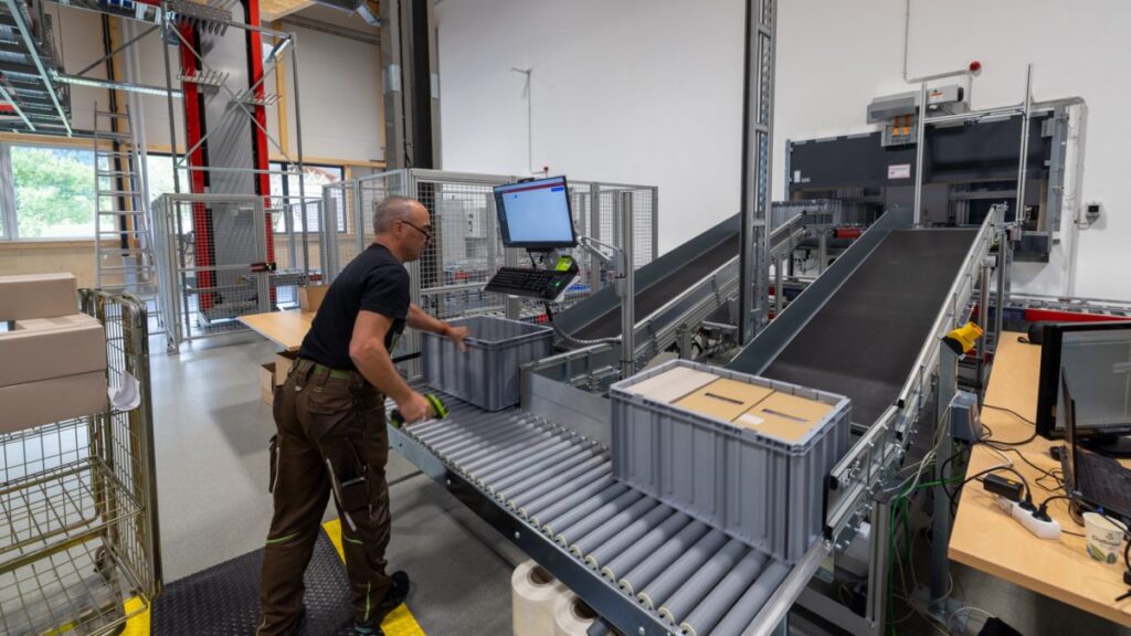 Un operario de almacén trabaja junto a un sistema de cinta transportadora automatizada en un almacén, utilizando una terminal informática para gestionar el flujo de trabajo, en línea con las estrategias de automatización 3PL para reducir costes laborales.