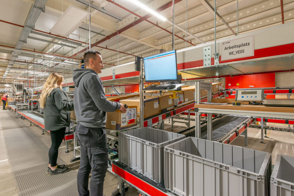 Operadores de FIEGE trabajando en estaciones de picking automatizadas con pantallas de control en el almacén.